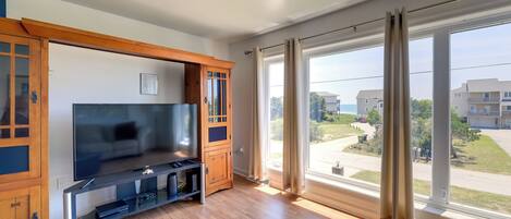 A modern living room with a large flat-screen TV, wooden cabinets, and a large window with a view of a residential area.
