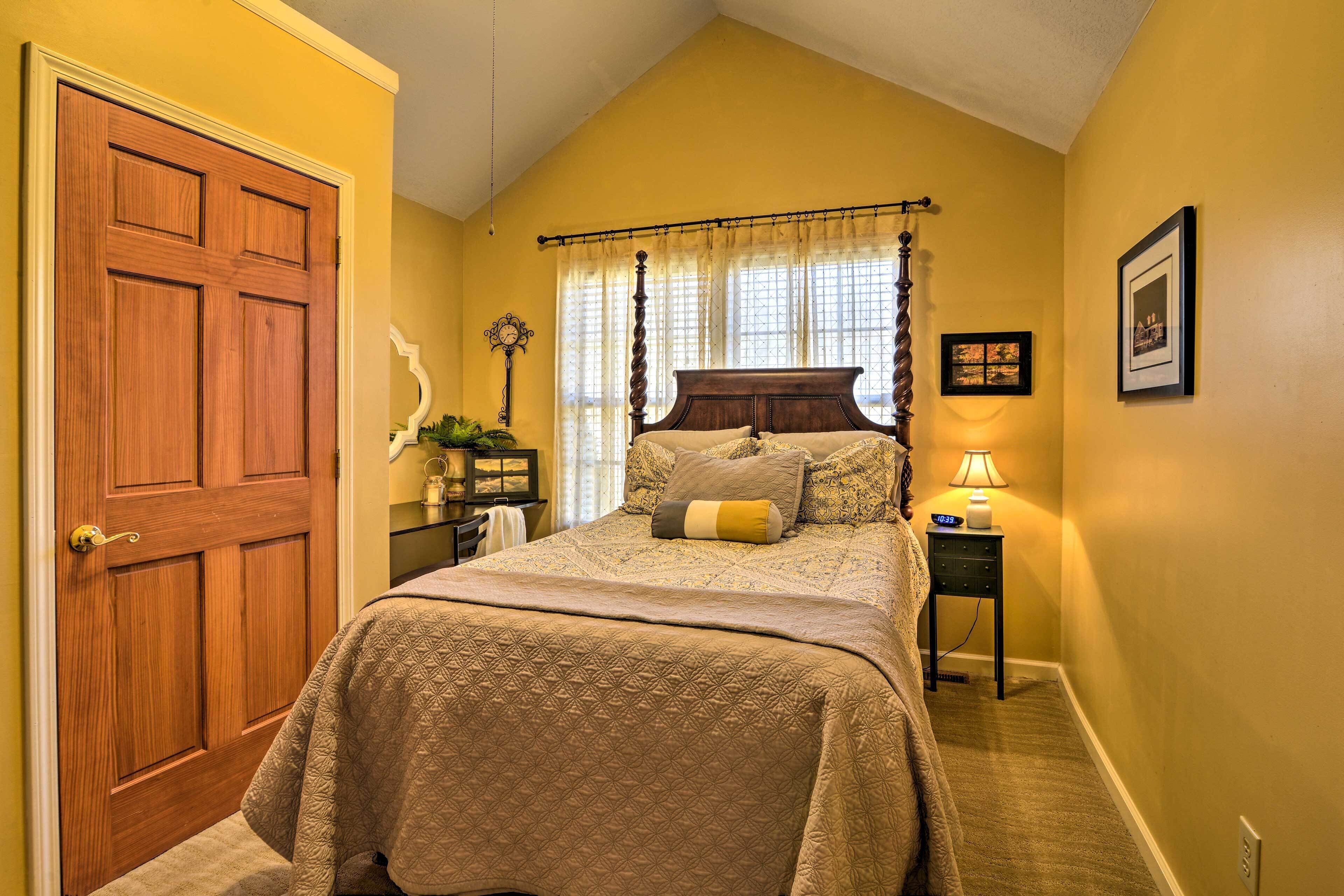 Quarto com porta de madeira, cama com colcha estampada, mesa de cabeceira com abajur e quadro pendurado na parede.