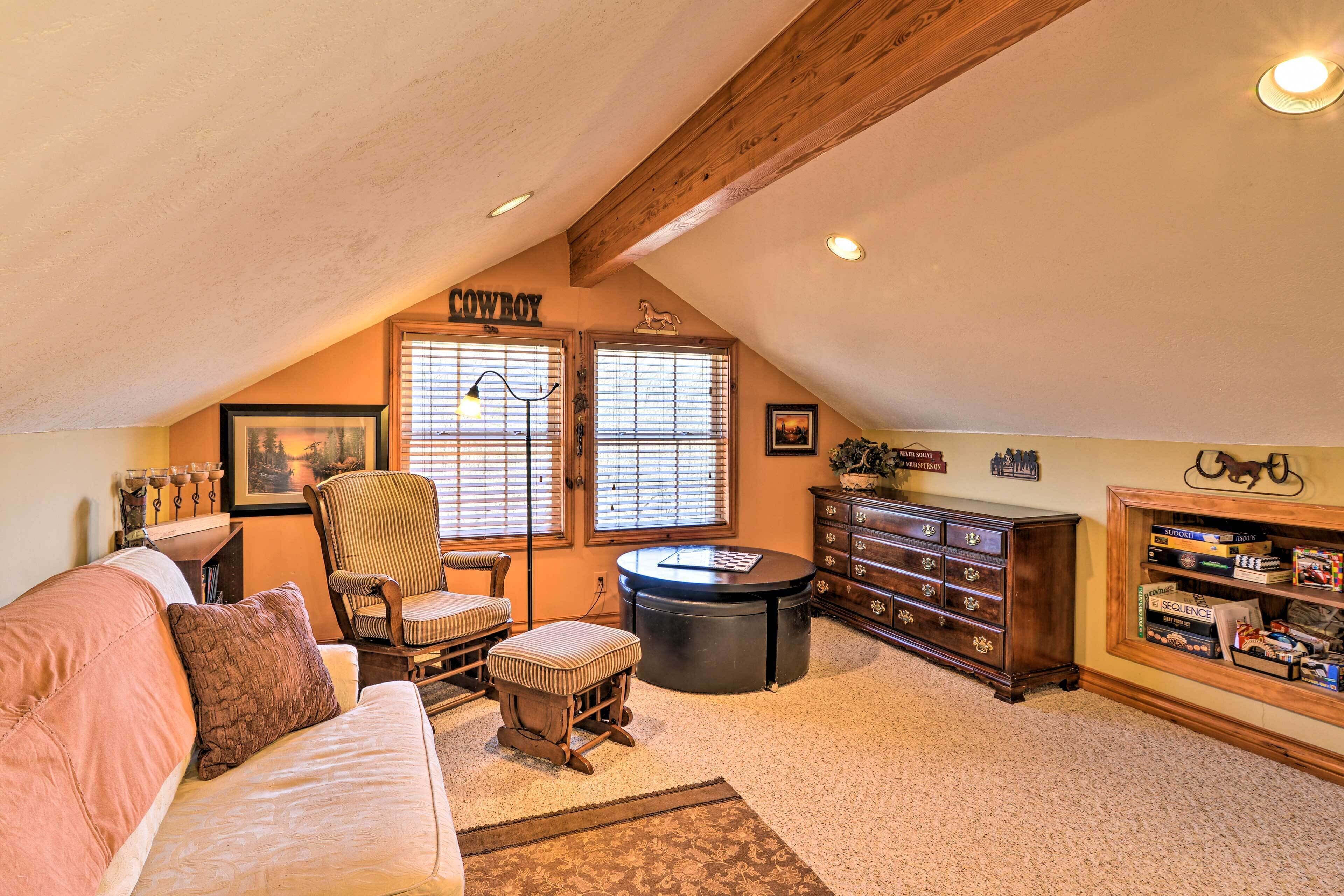 A cozy attic room with a high ceiling, a wooden ceiling beam, a sofa, a chair, a small table, a cabinet with board games, and a window with blinds.