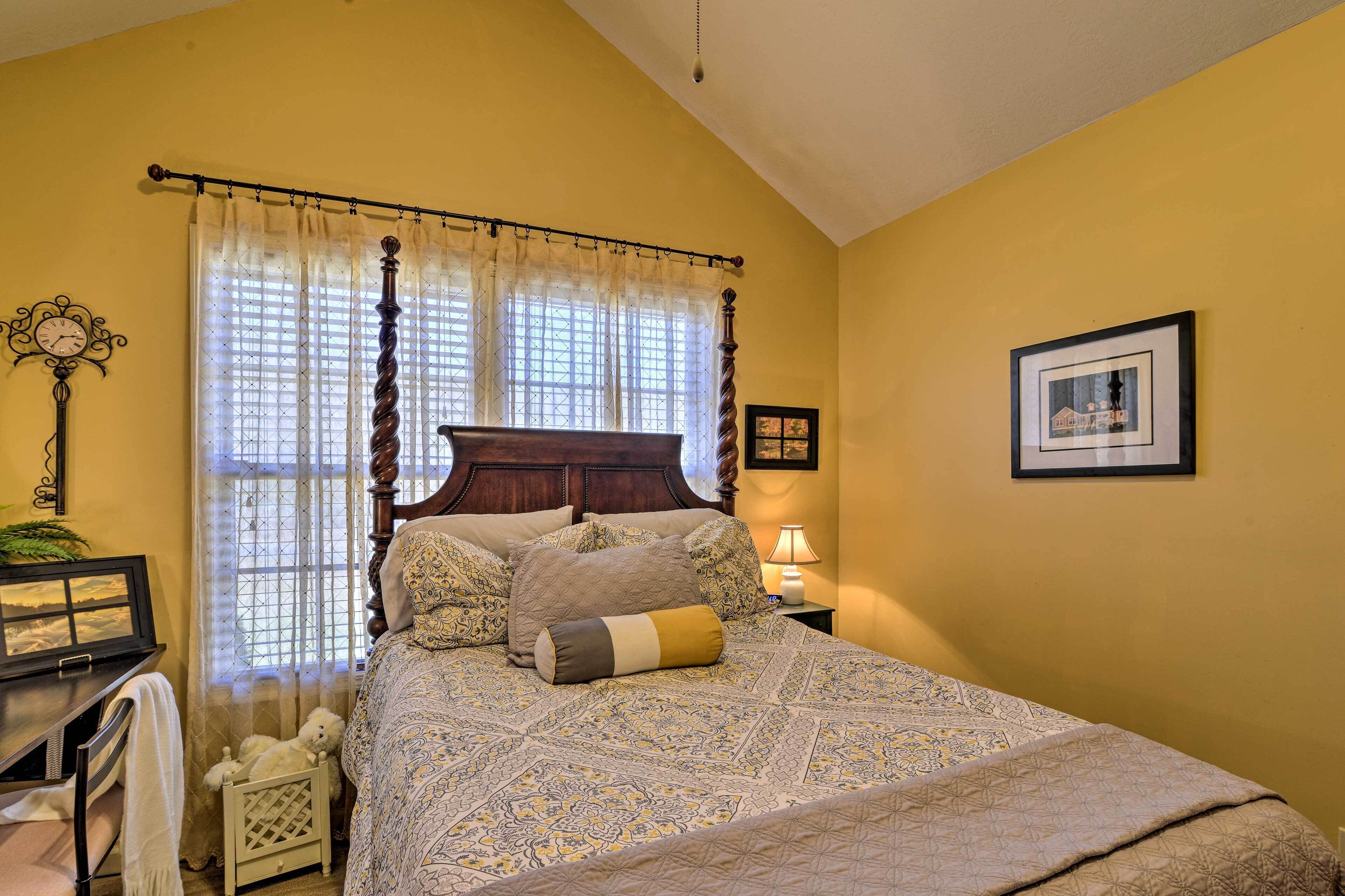 A bedroom with a bed, curtains, a clock, a framed picture, and a small table with a lamp.
