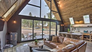 A spacious living area with a large leather sofa, a round coffee table, and a view of a forest through large windows. - Modern Lead A-frame w/ Hot Tub: Hike, Bike & Atv! (Lead)