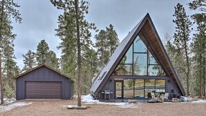 A modern A-frame cabin with a large glass window, a garage, and a gravel driveway surrounded by trees. - Modern Lead A-frame w/ Hot Tub: Hike, Bike & Atv! (Lead)