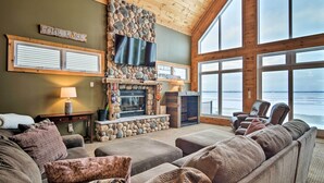 A cozy living room with a stone fireplace, comfortable seating, a flat-screen TV, and large windows overlooking a body of water.