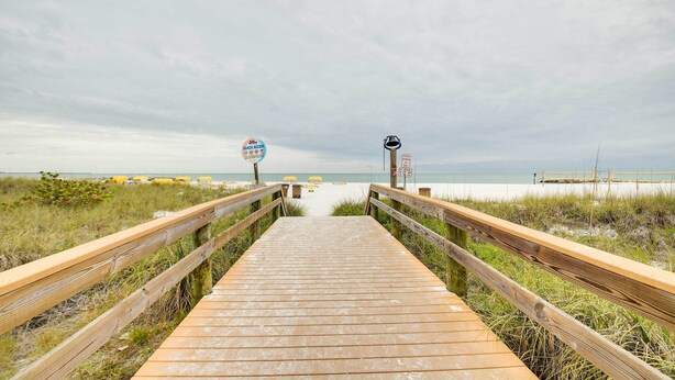 Ein Holzsteg führt zu einem Strand, wobei ein Schild den Zugang zum Strand kennzeichnet.