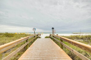 Une promenade en bois mène à une plage, et un panneau indique l’accès à la plage.