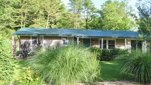 A single-story house with a green metal roof, surrounded by lush greenery and trees.
