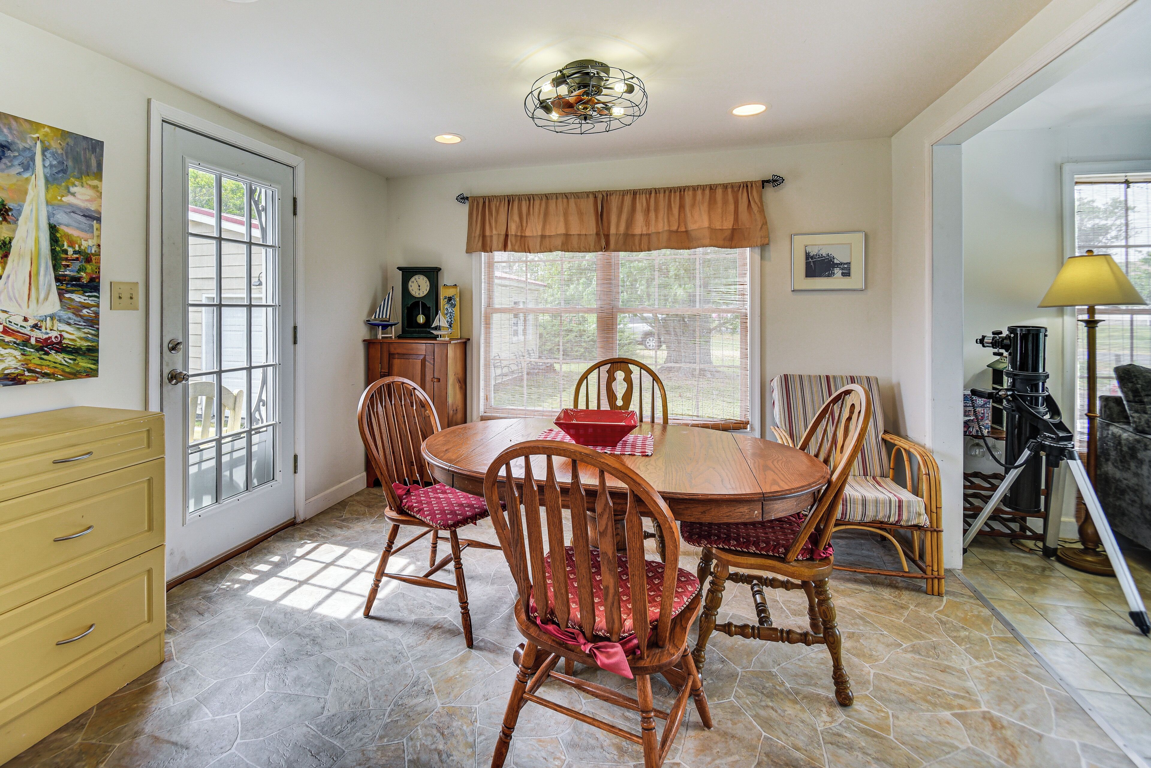 A dining room with a round table, four wooden chairs, a clock, and a window with a view.