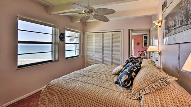 Un dormitorio con cama, ventilador de techo, ventana con vista al océano y puerta que da a otra habitación.