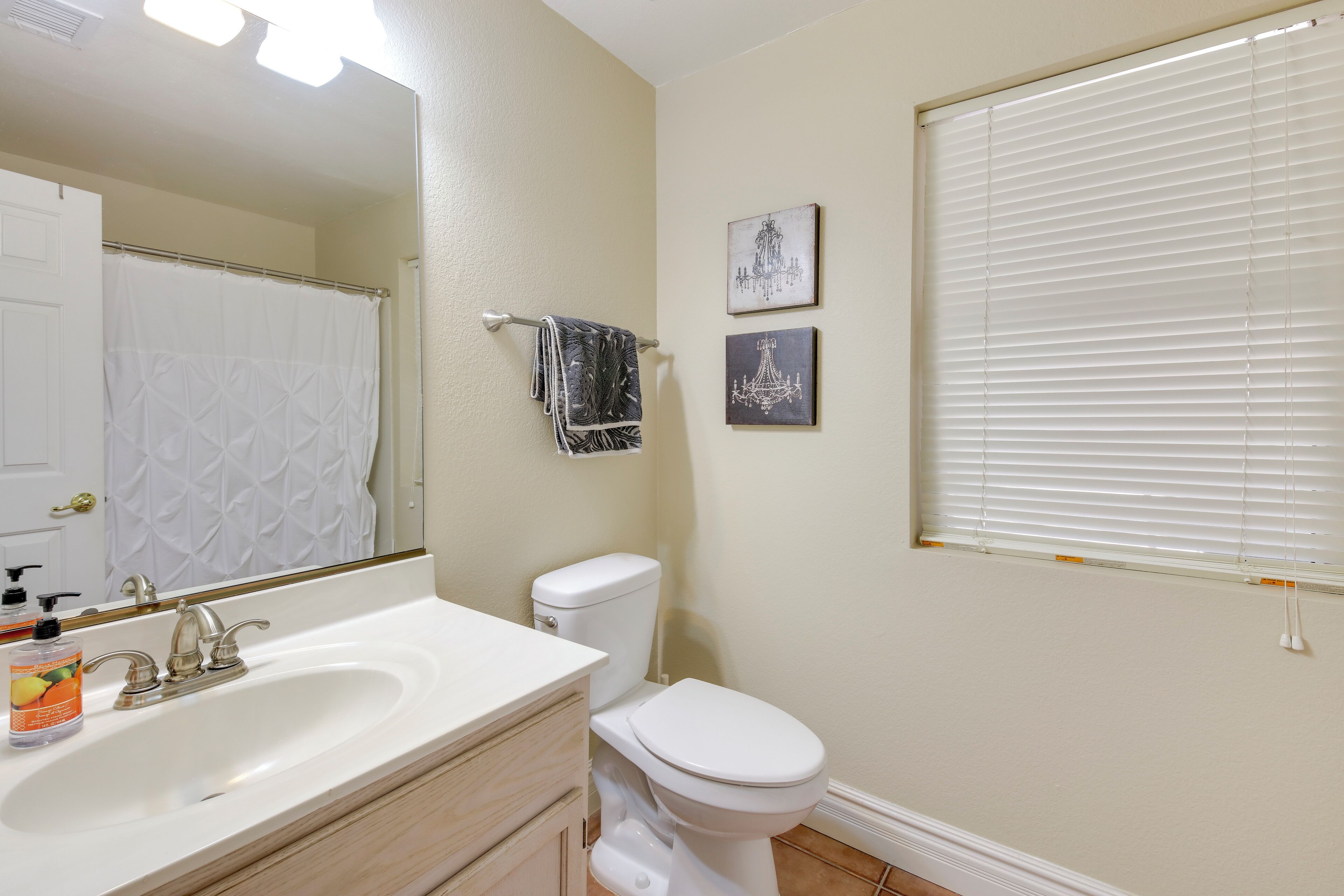A bathroom with a white sink, toilet, and a large mirror. There are two framed pictures on the wall and a window with blinds.