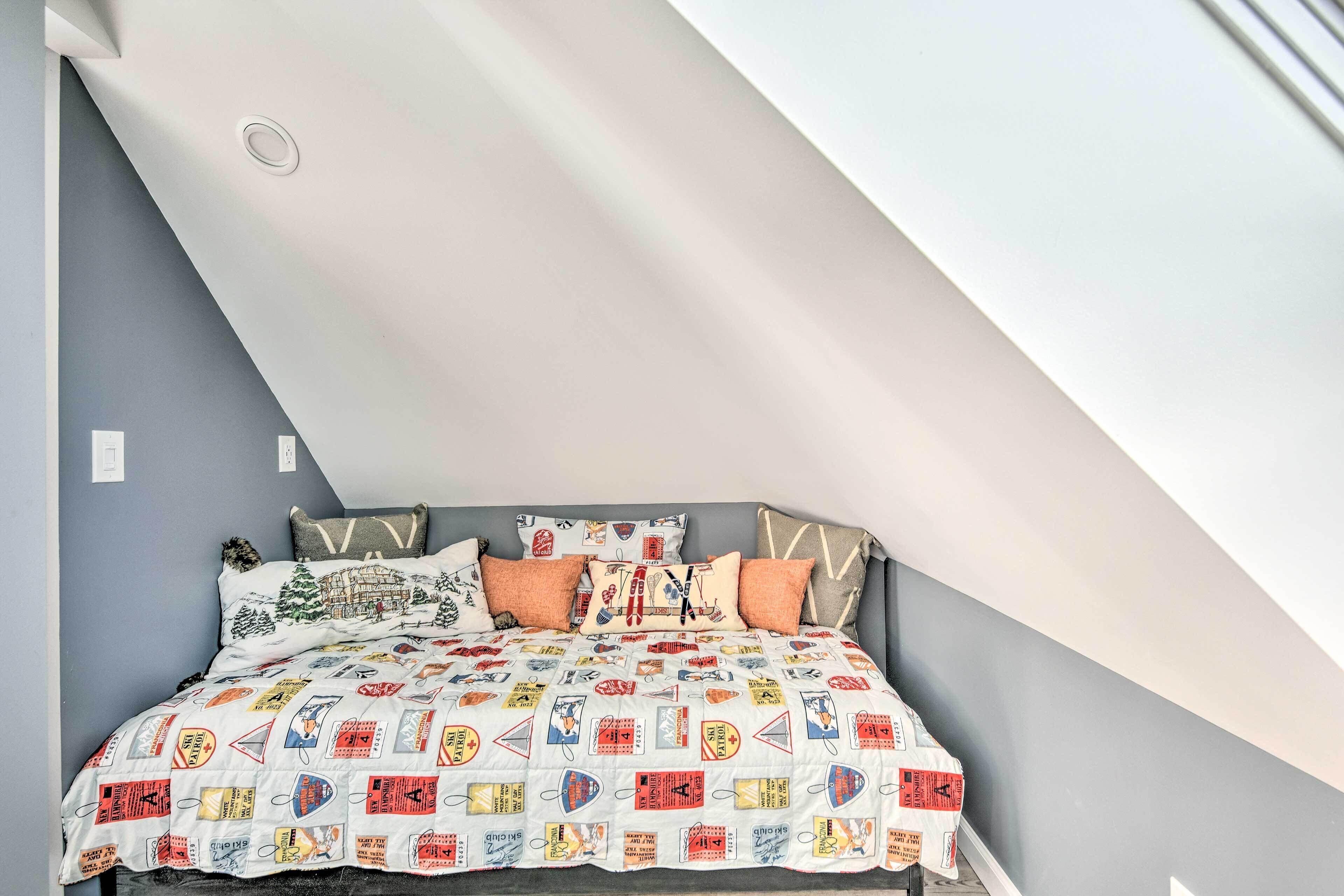 Une chambre avec un plafond en pente, un lit recouvert d’une courtepointe à motifs et une tête de lit grise.