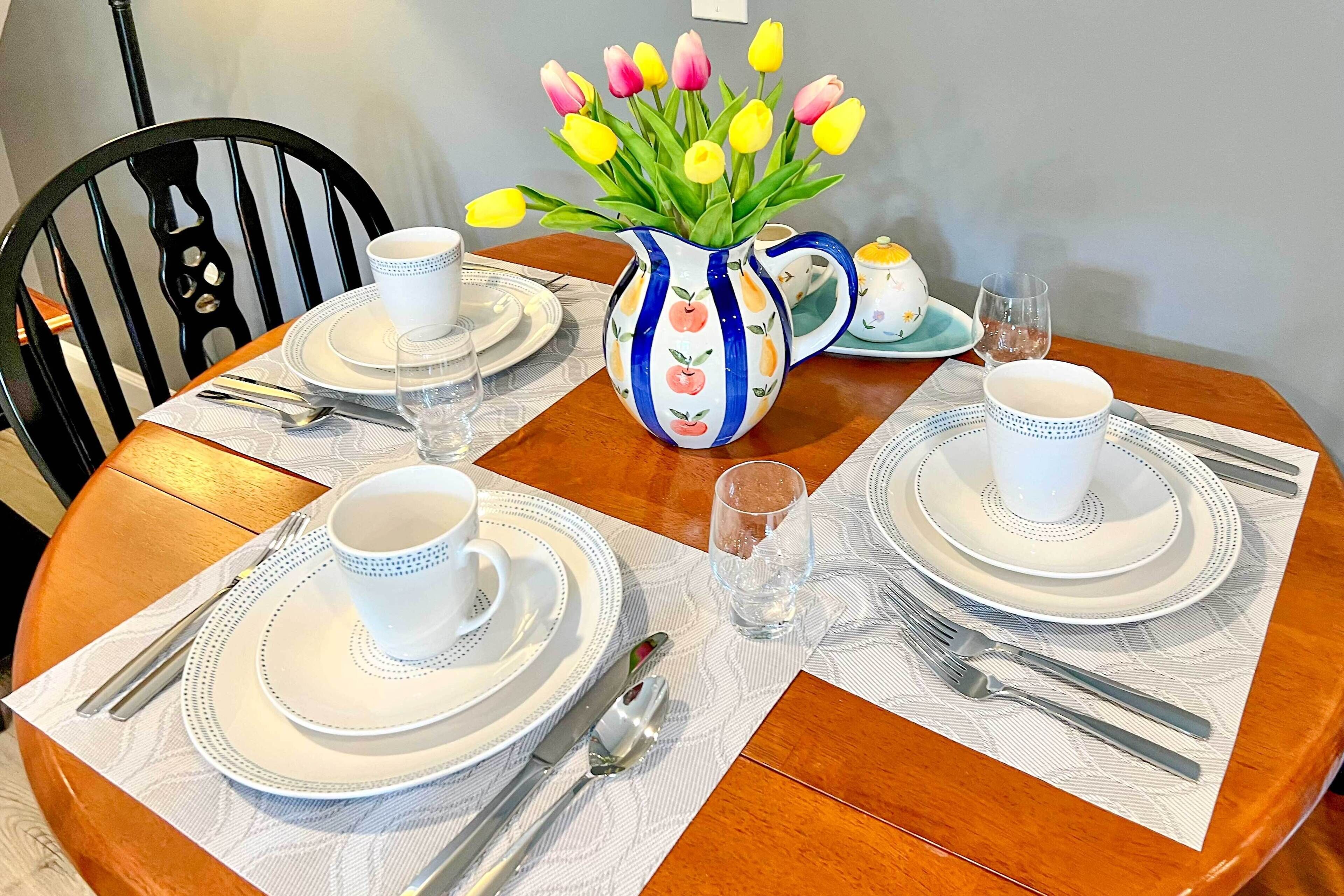 A dining table set with white plates, cups, and cutlery, a pitcher with tulips, and a black chair.