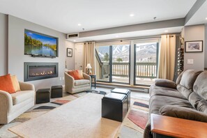 A living room with a fireplace, sofas, a coffee table, and a view of a mountain landscape through large sliding doors. - Walk to Lifts & Village! Killington Mountain Condo (Killington)