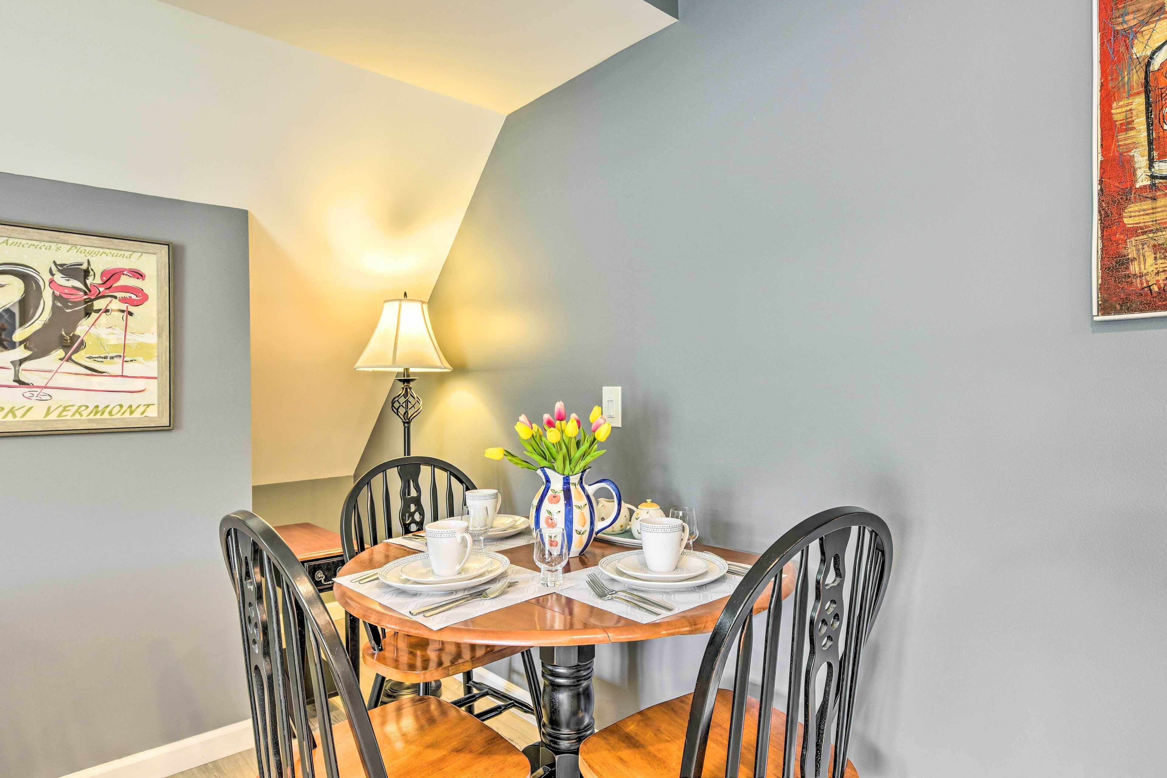 Un coin repas avec une table ronde dressée pour quatre personnes, entourée de chaises noires. Un vase rempli de fleurs jaunes est posé sur la table, et une lampe se trouve à l’arrière-plan.