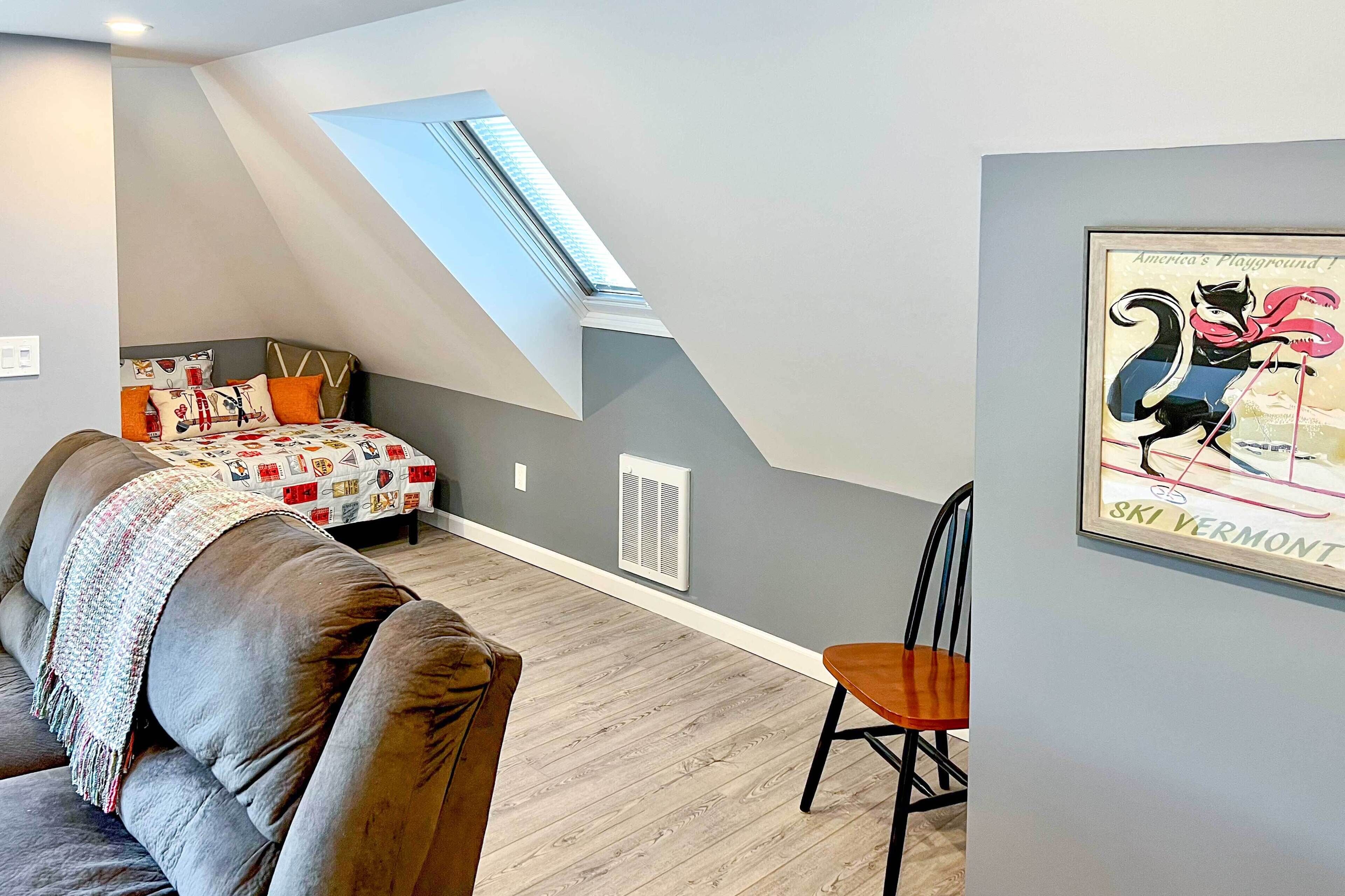 A bedroom with a sloped ceiling, a skylight, a bed with patterned bedding, a chair, and a framed poster on the wall.