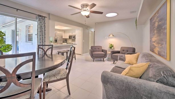 A modern living room with a grey sofa, a dining table with chairs, a ceiling fan, and a kitchen in the background.