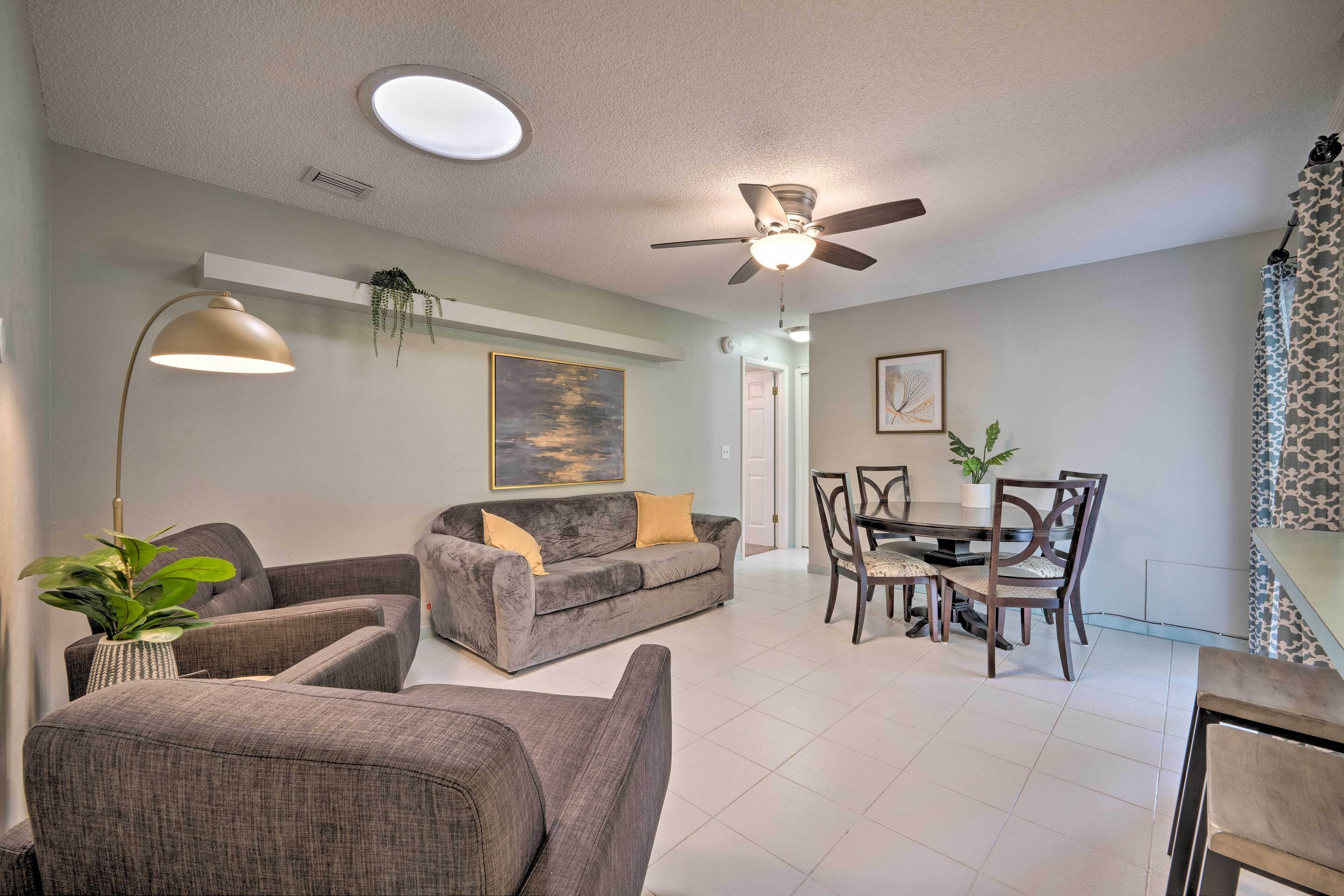 A living room with a gray sofa, two armchairs, a dining table with chairs, a ceiling fan, recessed lighting, and a potted plant.