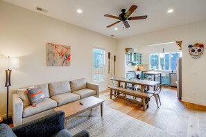 A spacious living area with a sofa, coffee table, and a dining table with chairs. There is a ceiling fan, artwork on the wall, and a view into the kitchen area.