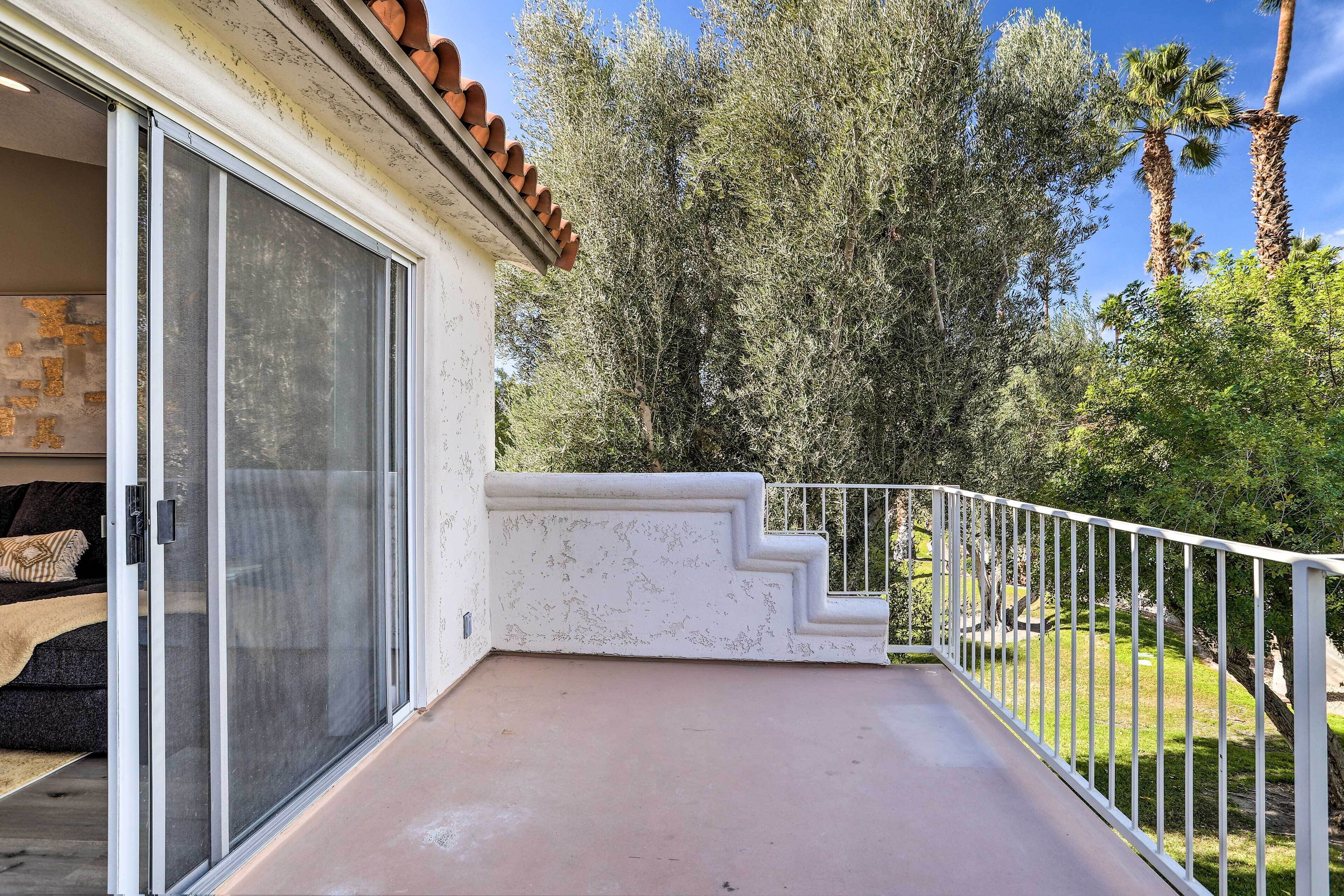 A balcony with a white railing, a sliding glass door, and a view of a garden with trees and a palm tree.
