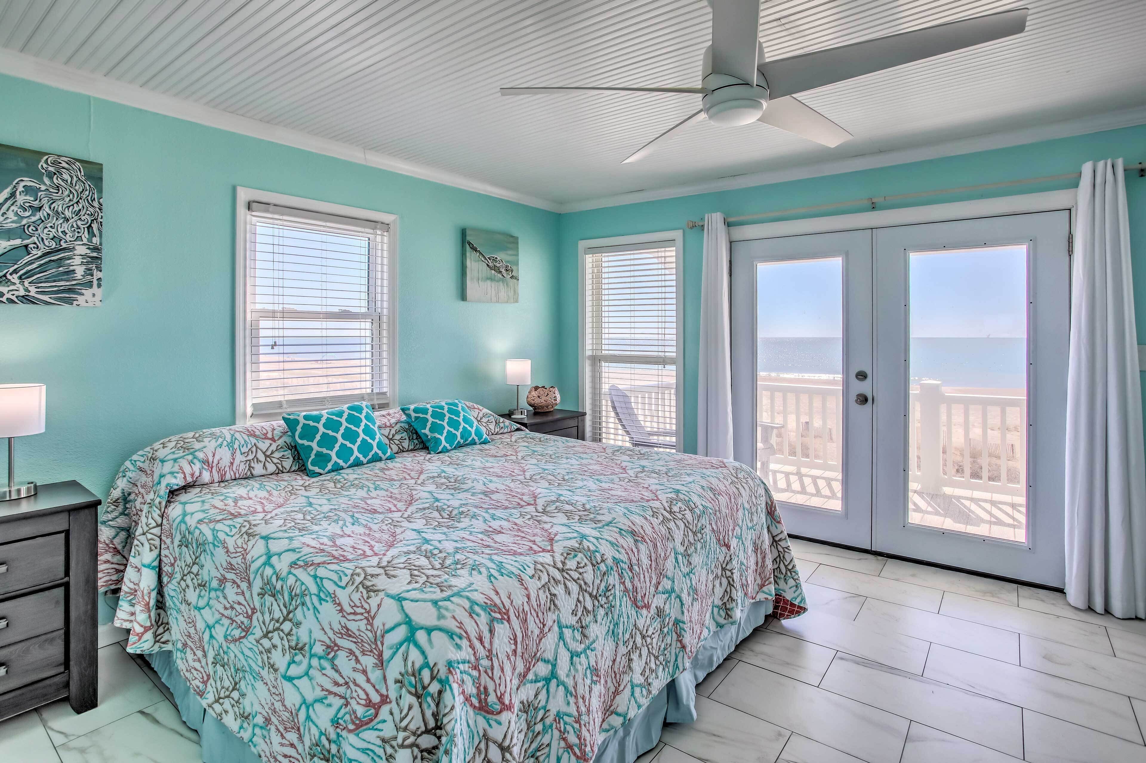 Quarto com cama, mesa de cabeceira, luminária, ventilador de teto e vista para o oceano pelas portas de vidro deslizantes.