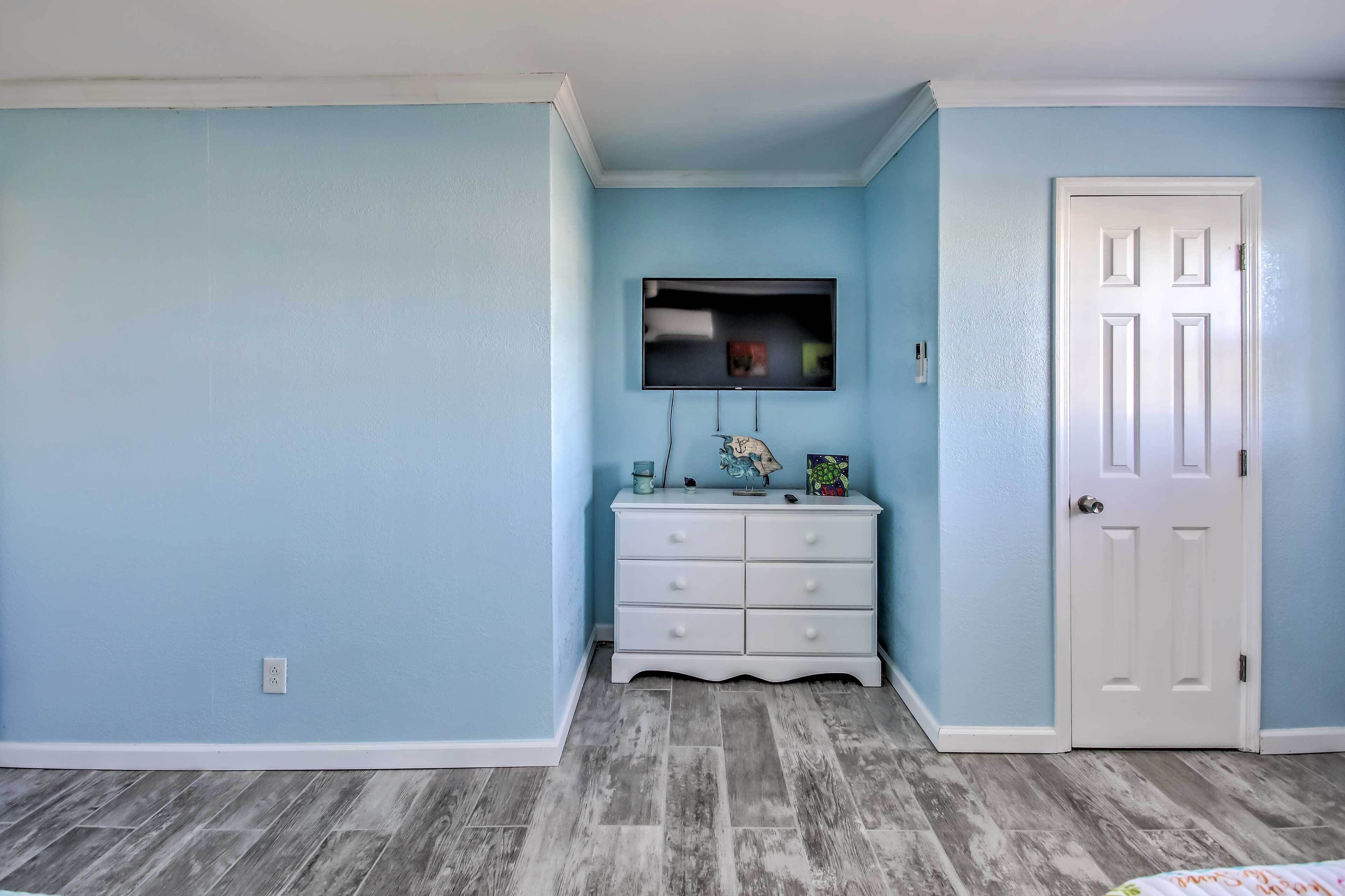 Quarto com paredes azul-claras, uma porta branca e uma cômoda branca com uma televisão de tela plana montada acima.