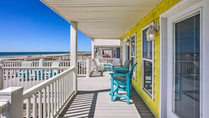 A covered porch with a view of the beach, featuring a white railing, blue chairs, and a yellow wall.
