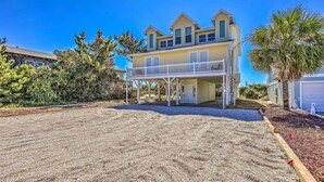 A two-story house with a balcony, surrounded by a sandy driveway and a small garden area.
