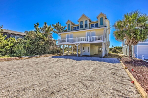 A two-story house with a balcony, surrounded by a sandy driveway and a small garden area.