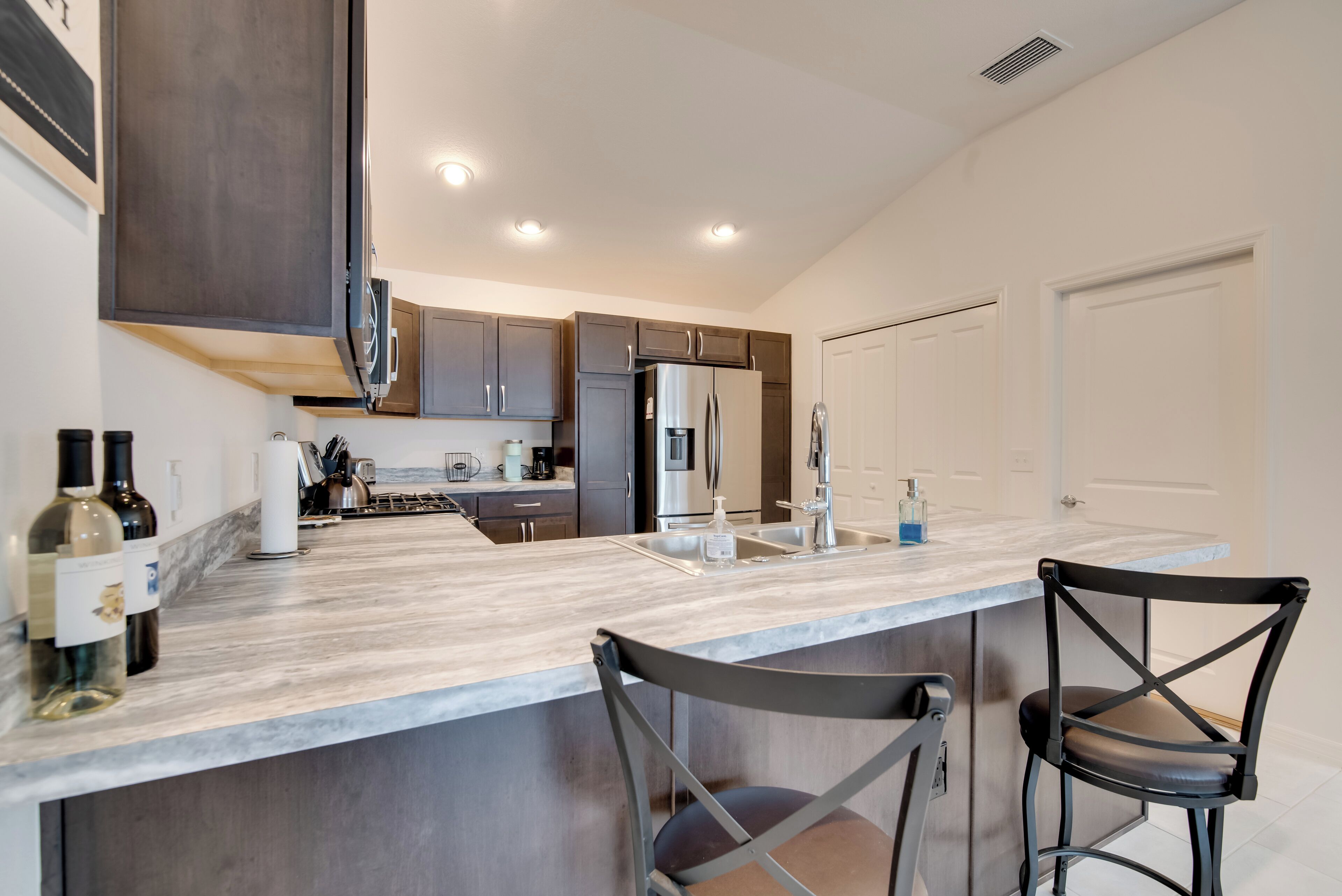 A modern kitchen with dark cabinets, a stainless steel refrigerator, a marble countertop, and two black bar stools.