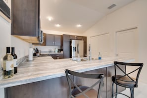 A modern kitchen with dark cabinets, a stainless steel refrigerator, a marble countertop, and two black bar stools.