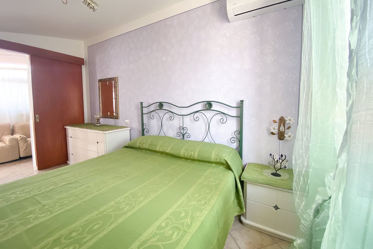 Un dormitorio con colcha verde, cómoda blanca, espejo y aire acondicionado.