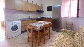 A compact kitchen with wooden cabinets, a washing machine, a dining table with chairs, a refrigerator, and a window with curtains. - Casa Vacanze da Vicenta (Camerota)