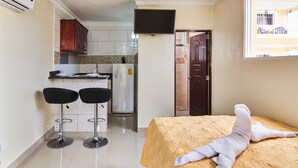 Une chambre d’hôtel compacte comprenant un lit, une petite kitchenette, deux tabourets de bar et un téléviseur fixé au mur.