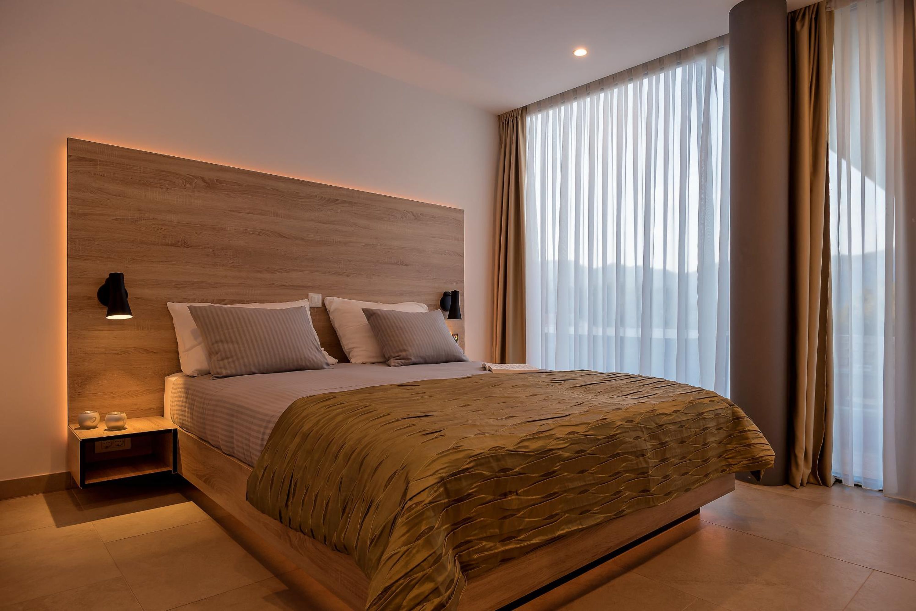 Un dormitorio moderno con una cama grande, una mesita de noche, una lámpara de pared y un amplio ventanal con cortinas.