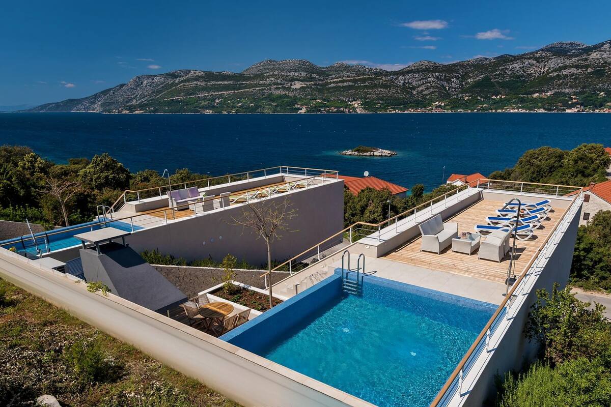 Una piscina en la azotea con sillones de descanso, un trampolín y vistas al mar y a las montañas.