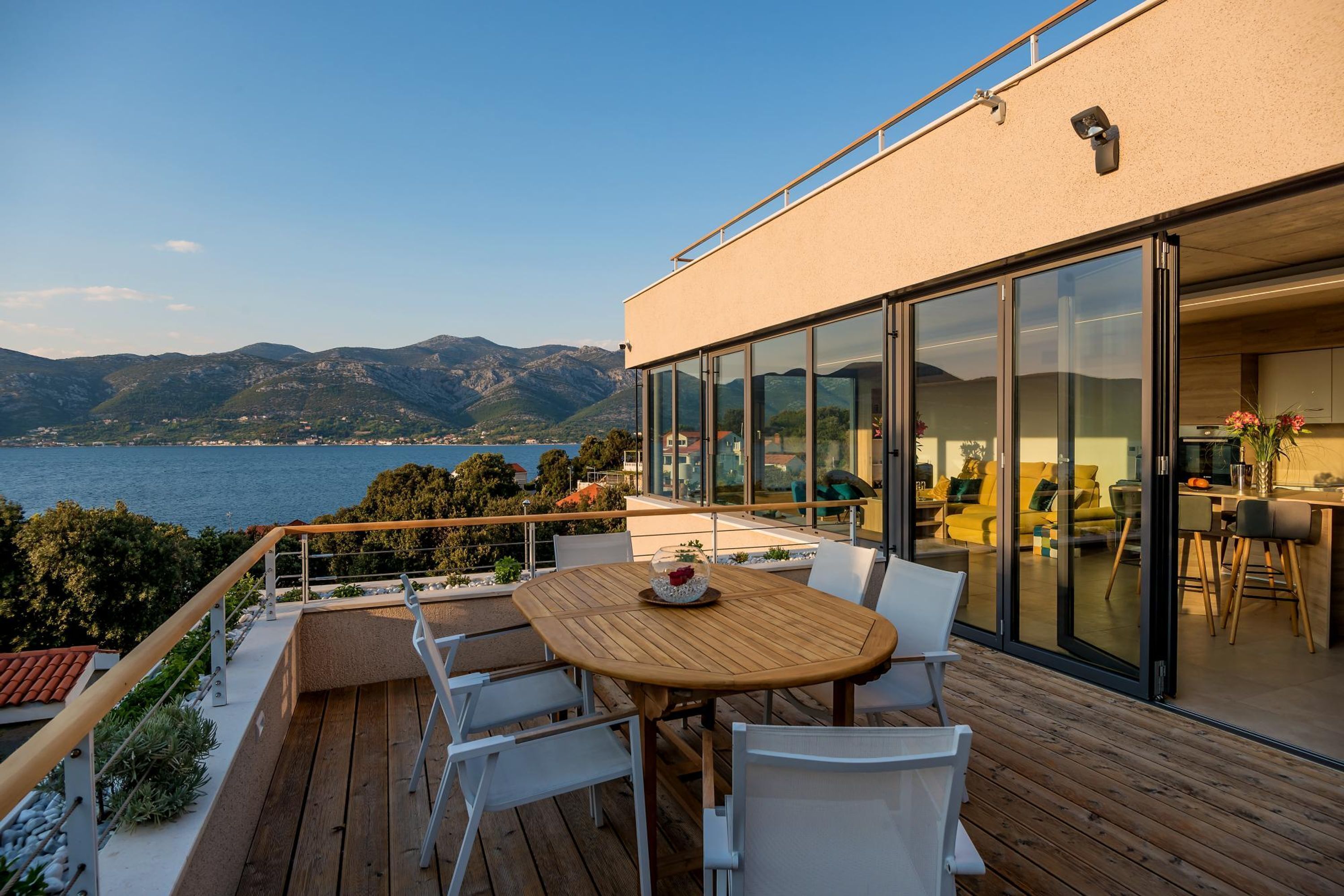 A modern outdoor deck with a wooden table and chairs, overlooking a body of water and mountains.
