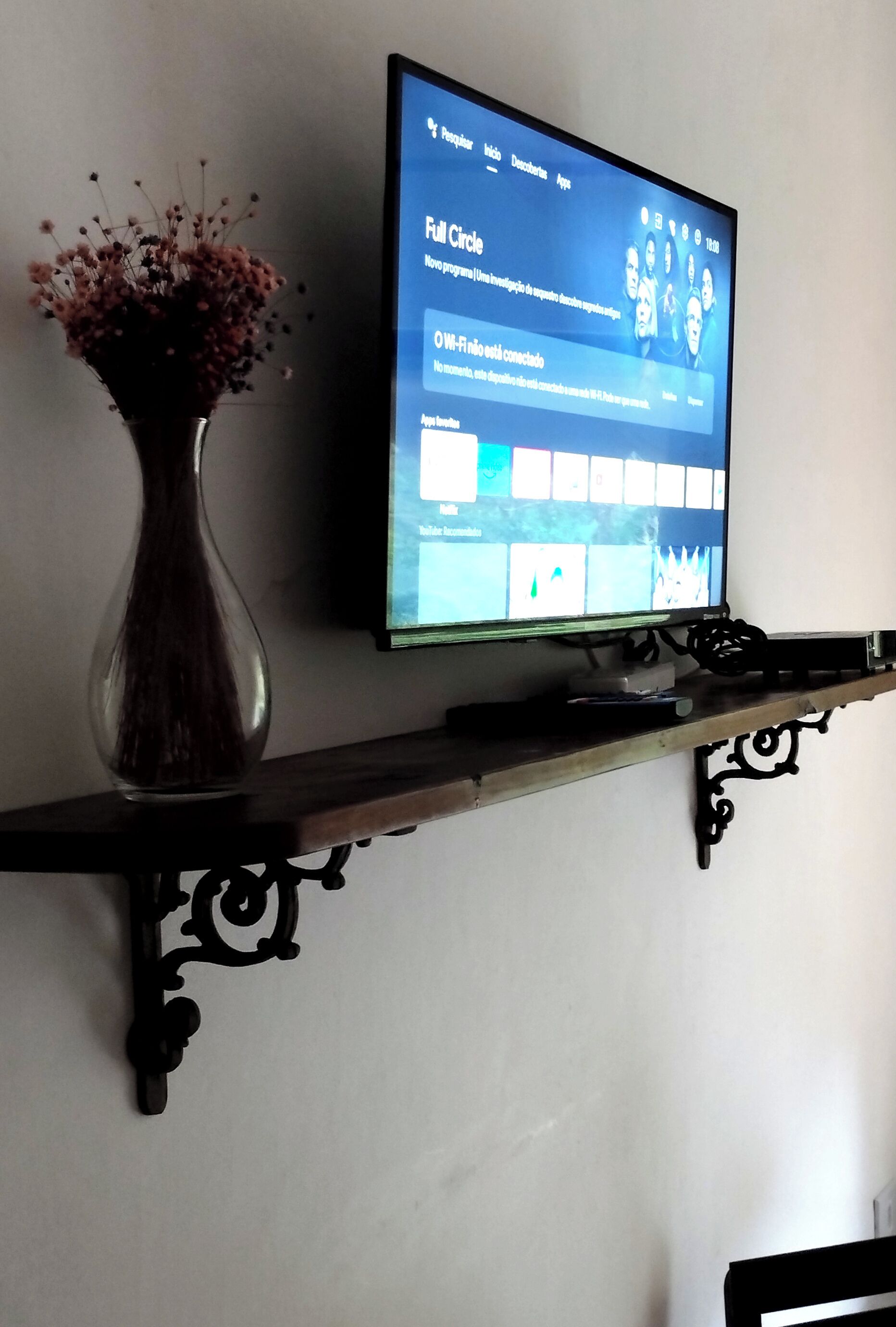 A television on a wall-mounted shelf with a vase of dried flowers to the left.