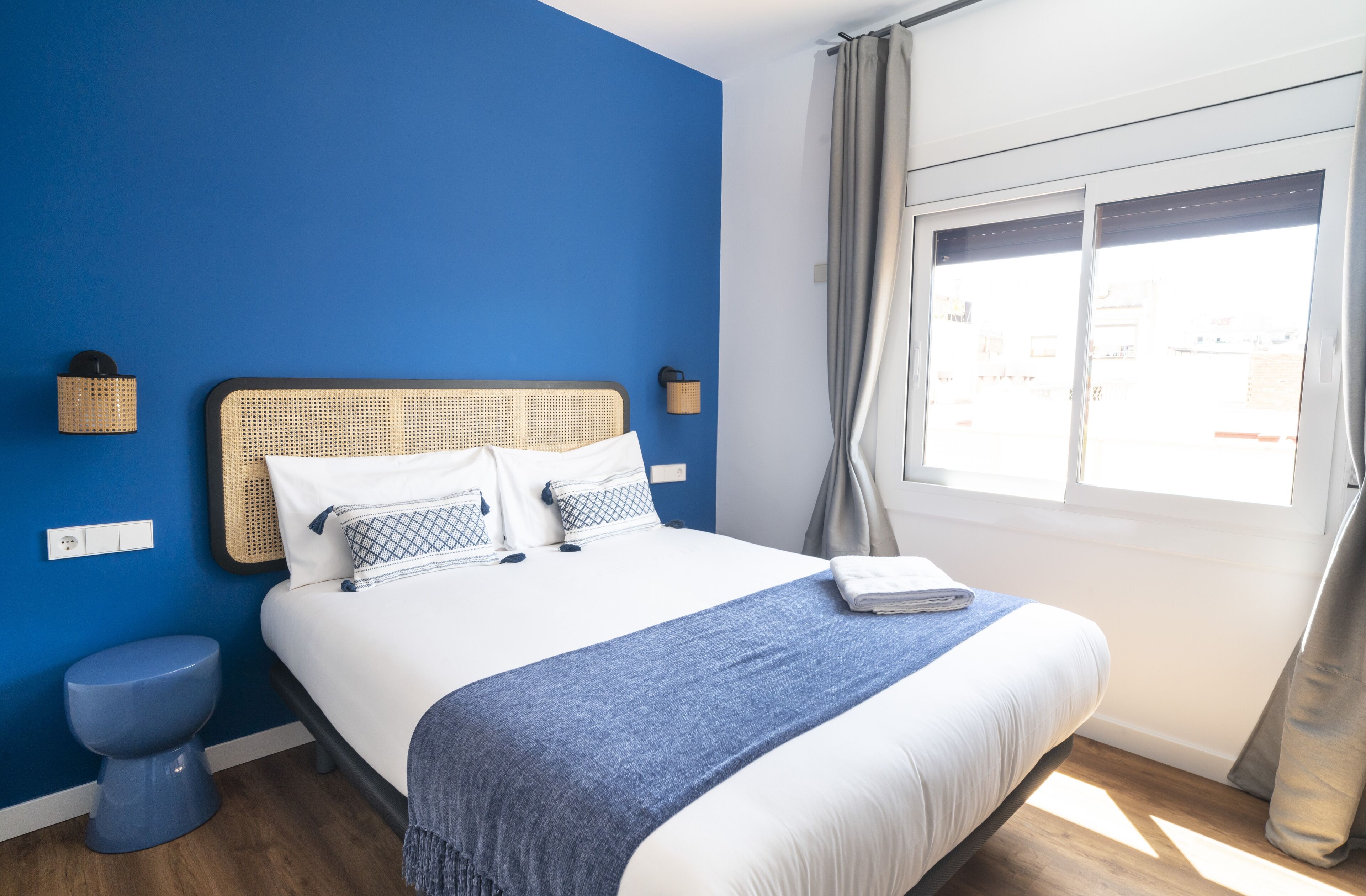 A hotel room with a large bed, a blue wall, a window with curtains, and a wooden floor.