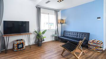 Una sala de estar con un sofá negro, una mesa de centro de madera, un televisor sobre un mueble, una planta en maceta y una lámpara.