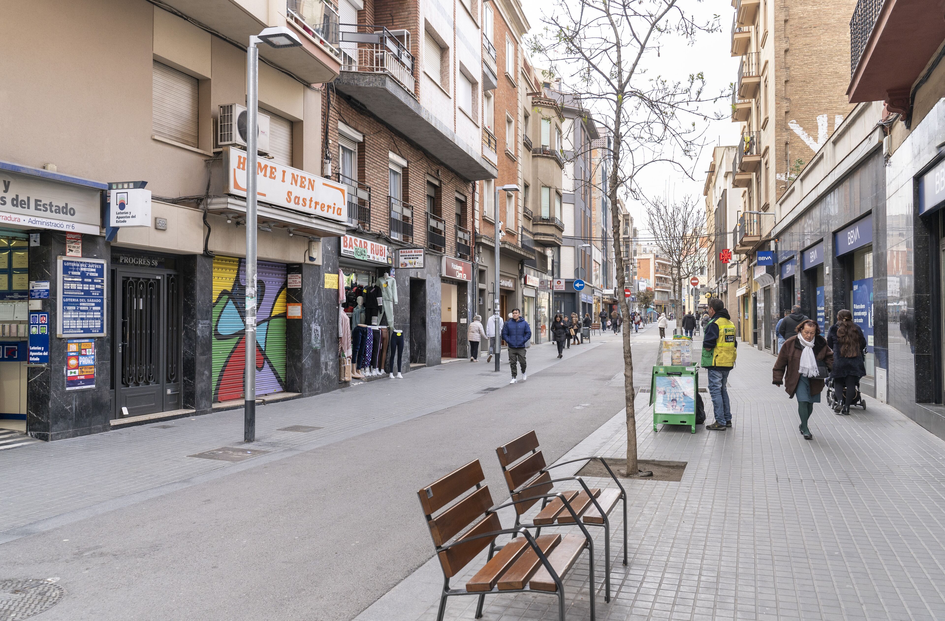 Una calle con bancos, tiendas y gente caminando.