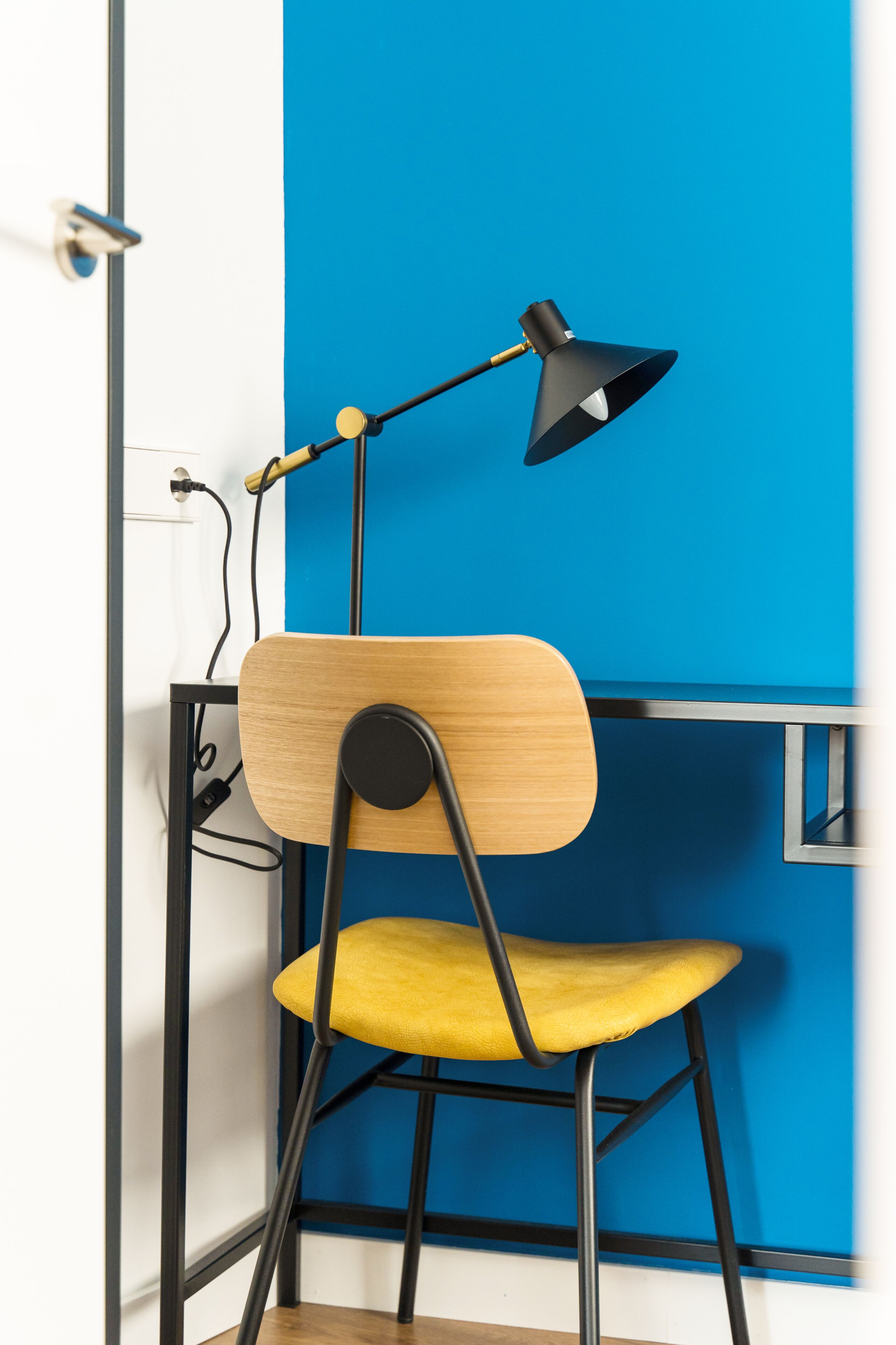 Una silla con respaldo de madera y asiento amarillo, un escritorio negro y una lámpara de escritorio negra.
