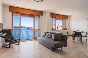 Un salon moderne avec un canapé gris, un fauteuil noir et une table à manger avec des chaises. Une grande fenêtre offre une vue sur la mer et les bâtiments.
