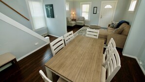 A dining area with a wooden table and white chairs, a staircase, a living room with a sofa and a lamp, and a hallway with a door.