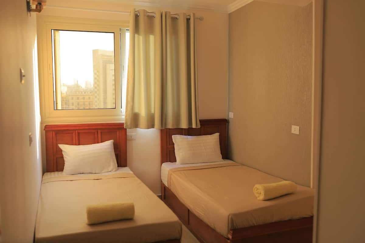 A room with two single beds, a window with curtains, and a view of buildings outside.