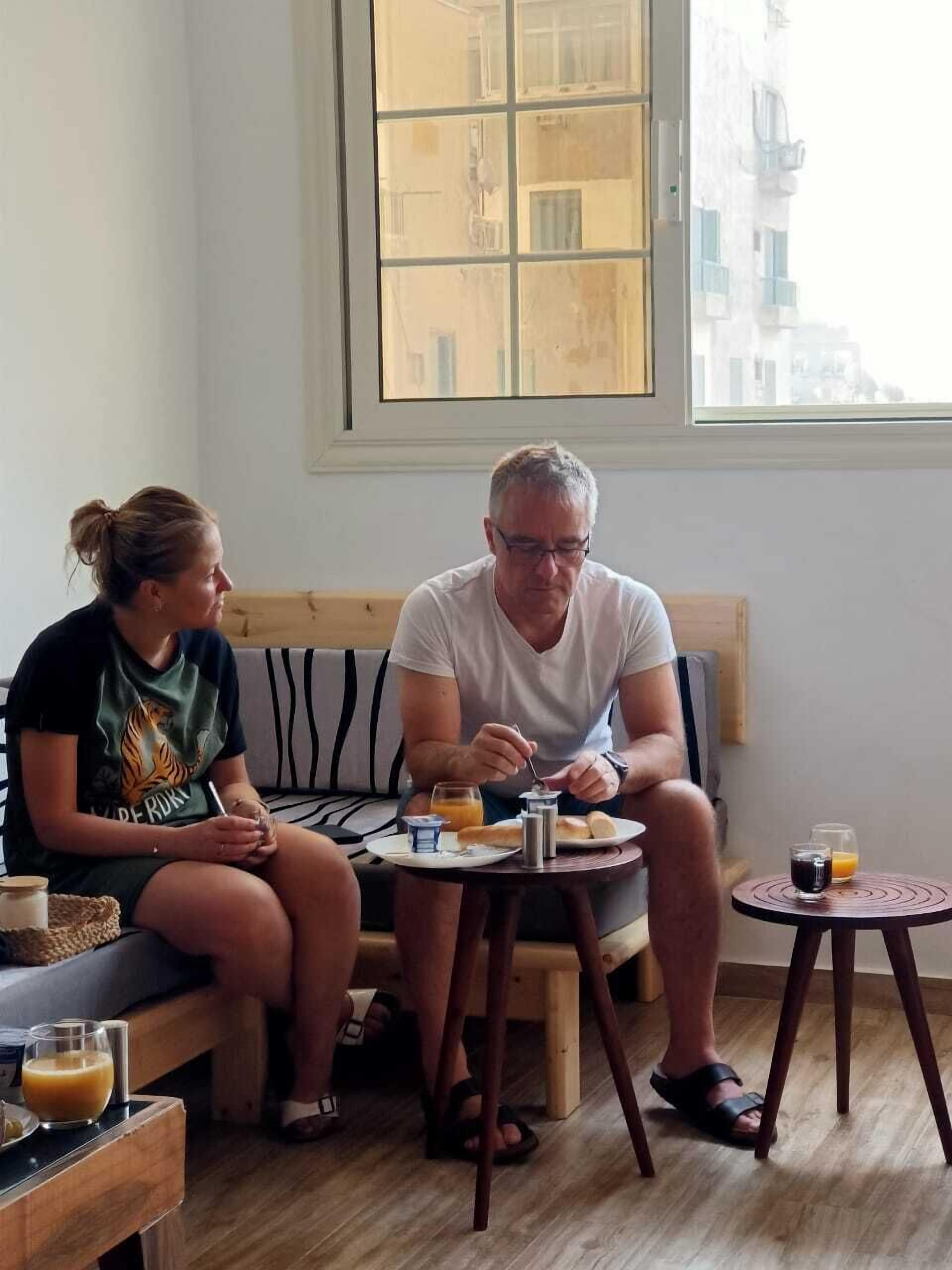 Una habitación con un banco de madera, una pequeña mesa redonda con cosas para el desayuno y una ventana que da a edificios.