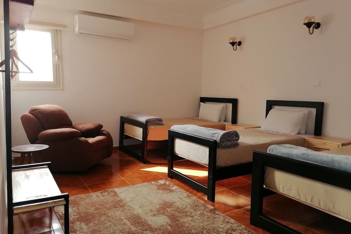 A small, well-lit room with two beds, a chair, a small table, and an air conditioning unit.
