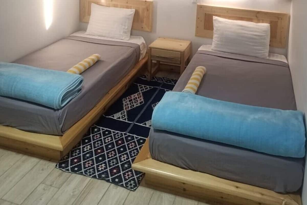A small, compact room with two single beds, a small wooden nightstand, a patterned rug, and a window with curtains.