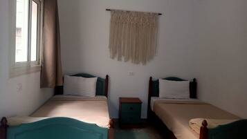 Una habitación con dos camas individuales, una mesita de noche pequeña, una cortina de macramé en la pared y una ventana con cortinas.