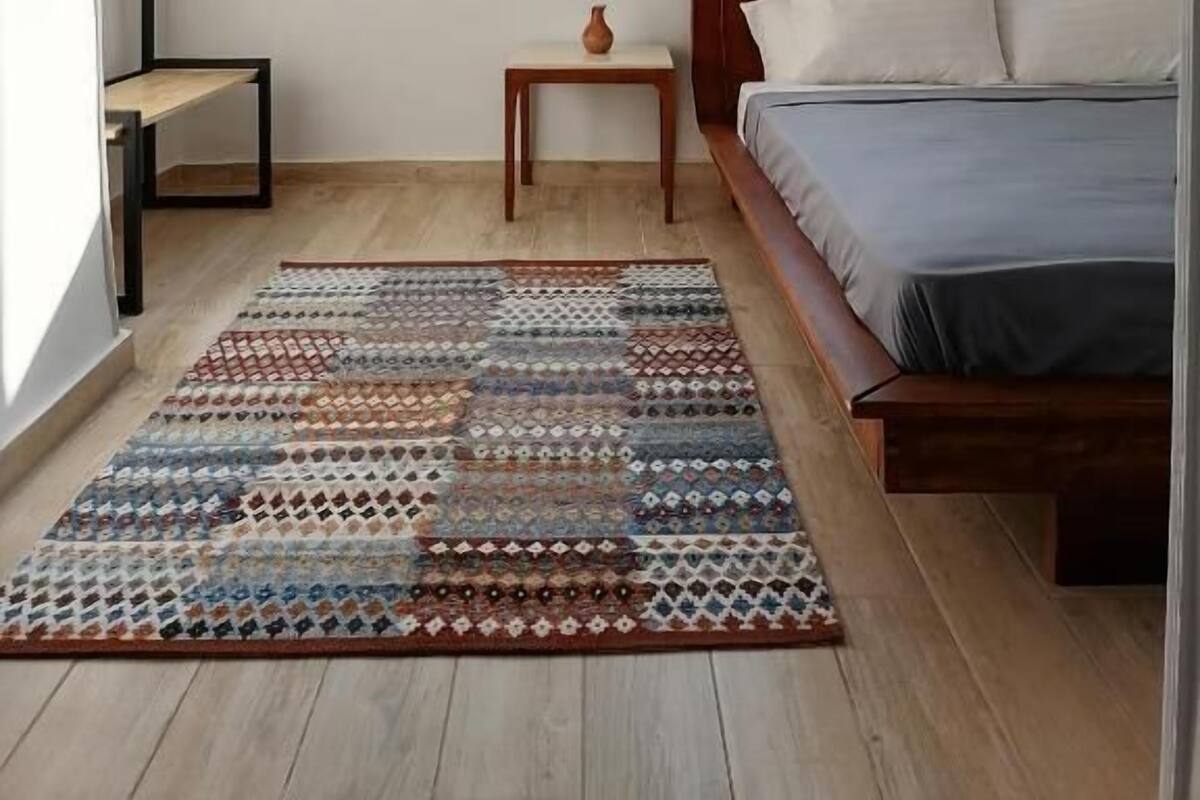 A bedroom with a bed, a window, a small table with a vase, and a patterned rug.