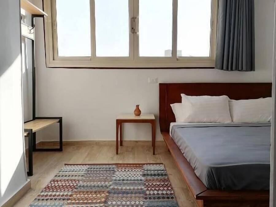 Un dormitorio con cama, una ventana, una mesita de noche con un florero y una alfombra estampada.
