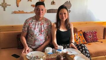 Una pareja sentada en un sofá frente a una mesa de madera, sosteniendo una bandeja con un plato, una taza y una cesta.